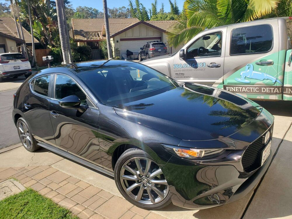 Black Mazda3 sedan freshly detailed in a driveway, with the Finetactic Detailing truck parked behind it